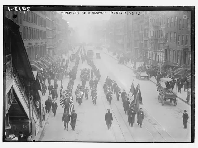 The image shows a black and white photo of a parade on a city street. There are many people walking...