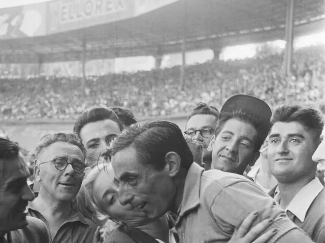 The image shows a black and white photo of a man embracing a woman in front of a crowd. The man is...