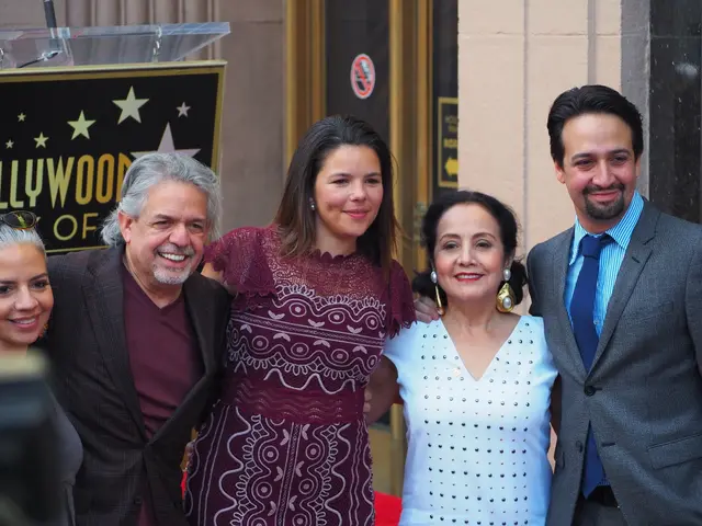 The image shows a group of people standing next to each other on a red carpet, smiling and posing...
