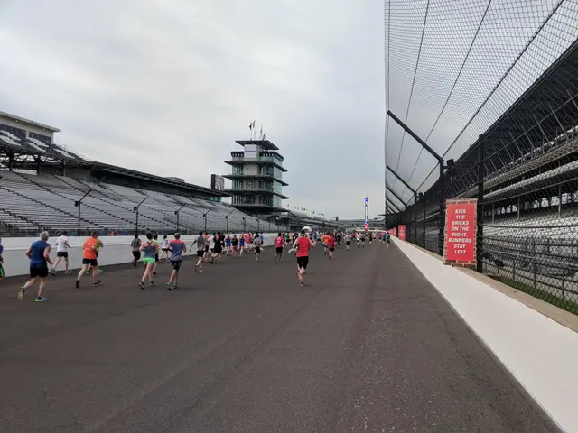 The image shows a group of people running down a race track, with a fence on the right side of the...