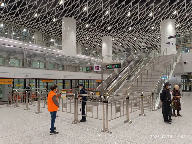 The image shows a group of people standing in an airport terminal with an escalator, railings,...