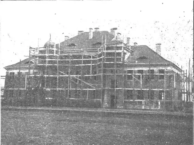 The image shows a large building under construction, with scaffolding around it. The photo is in...