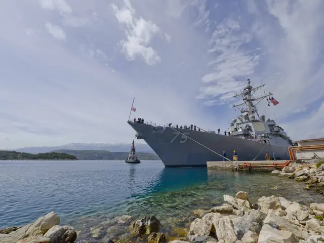 The image shows a navy ship docked at a dock in the water, surrounded by rocks and trees on the...