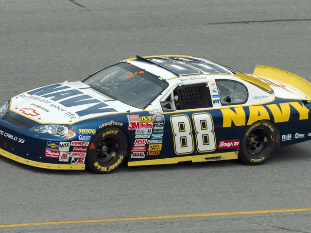The image shows a NASCAR car driving down a race track, with text on the car and grass at the...