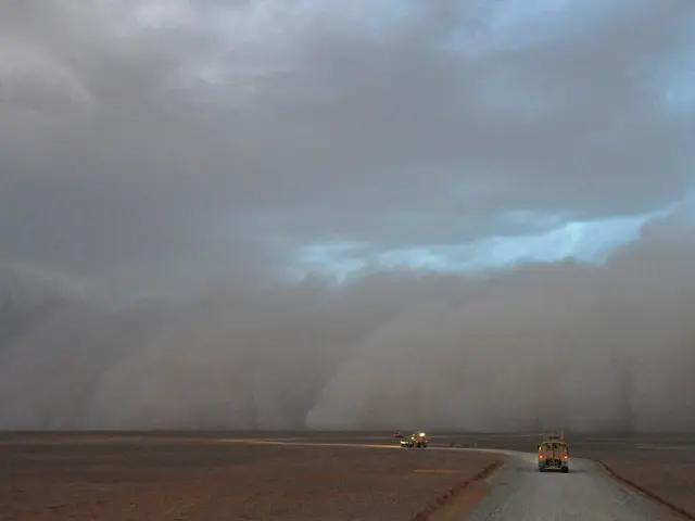 The image shows vehicles driving down a road in the middle of a desert, with a dust storm looming...