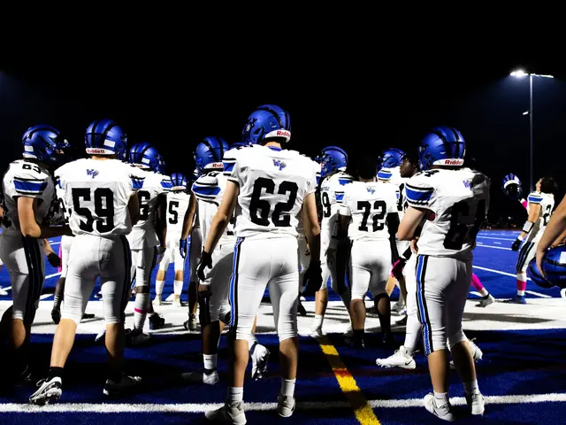 The image shows a group of football players standing on top of a field, illuminated by light poles...