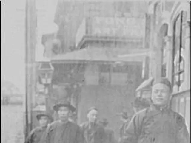 The image shows an old black and white photo of a group of people walking down a street, with...
