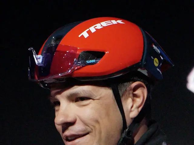The image shows a man wearing a red and white jersey and a helmet, holding a microphone in his...