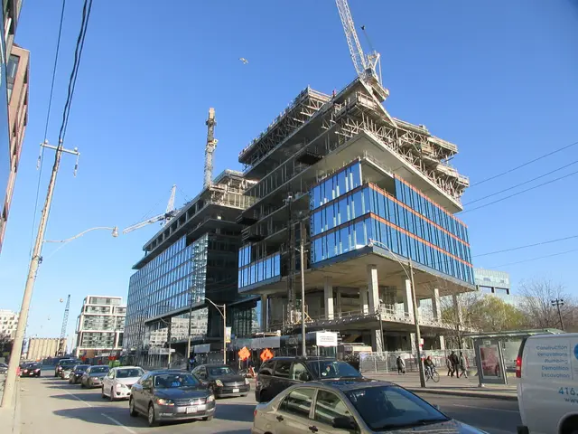 The image shows a large building under construction on the corner of a street, with vehicles...
