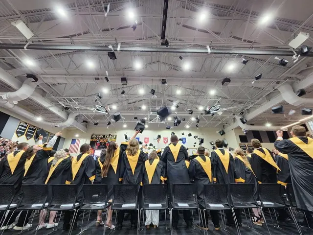 The image shows a group of people in graduation robes and hats standing in front of a crowd of...