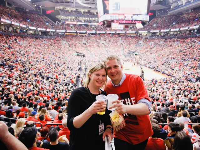 The image shows a man and woman standing in front of a large crowd in a stadium. They are both...