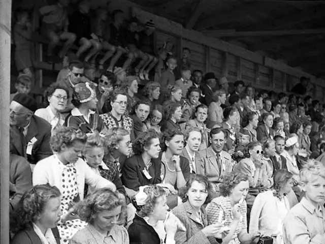 The image shows a black and white photo of a crowd of people sitting in a stadium, with a wooden...