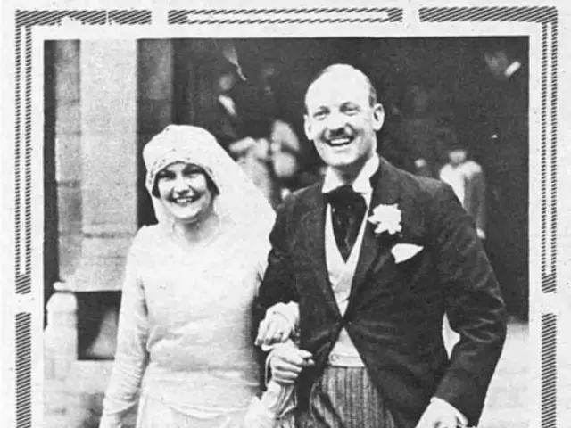 The image shows a black and white photograph of a man and woman walking down the street, the man...