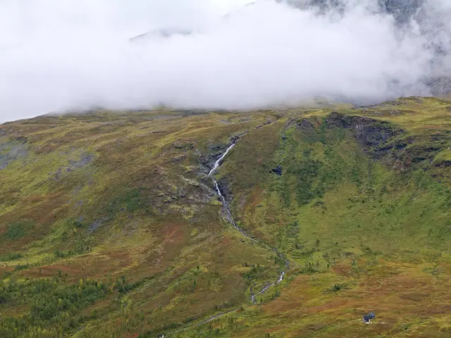 The image shows a small waterfall cascading down the side of a mountain, surrounded by lush green...