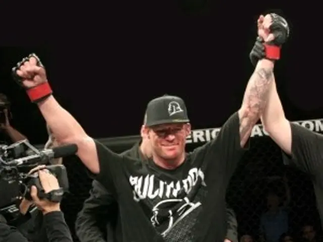 The image shows a man wearing a black t-shirt, black shorts, and a black cap, holding up a UFC belt...