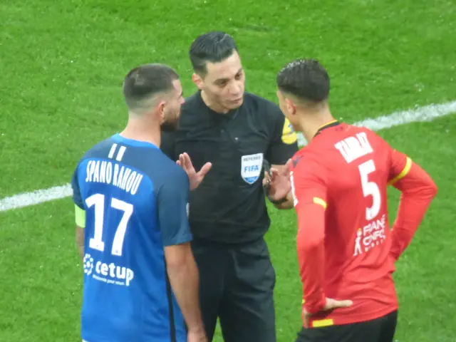 The image shows a referee talking to two men on a soccer field. The man in the middle is wearing a...