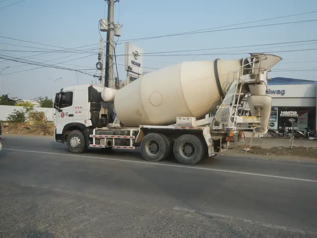The image shows a cement mixer truck driving down the street, surrounded by electric poles with...