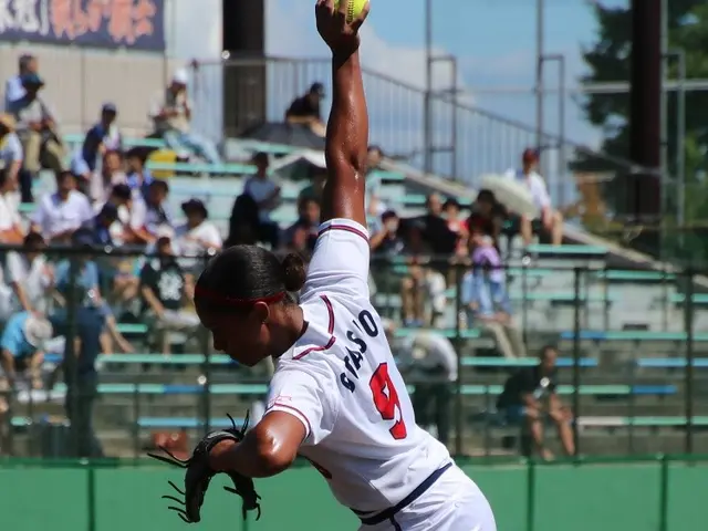 The image shows a woman in a white dress throwing a softball on top of a field. She is wearing a...
