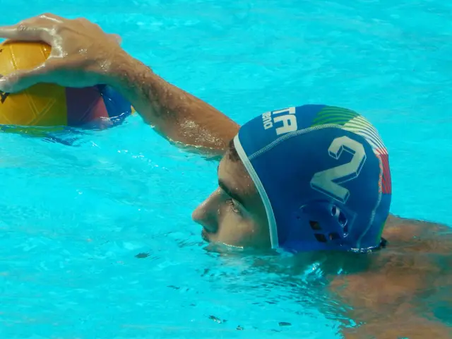 The image shows a water polo player in a pool, wearing a cap and holding a ball in his hand. He is...