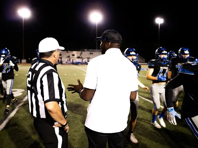 The image shows a football coach talking to his team on the field at night. The players are wearing...