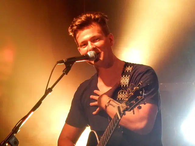 The image shows a man playing a guitar and singing into a microphone in front of a crowd. He is...