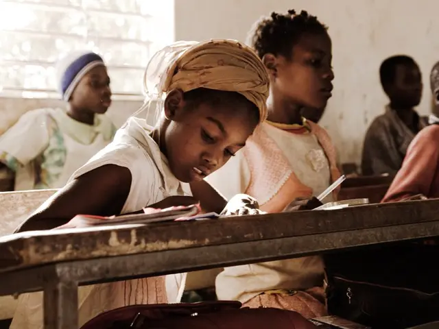 The image shows a group of children sitting at desks in a classroom, with books and bags on the...