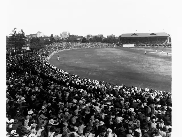 The image shows a black and white photo of a large crowd of people watching a cricket match at the...