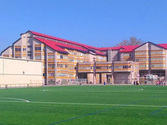 The image shows a soccer field with a large building in the background, surrounded by trees and...