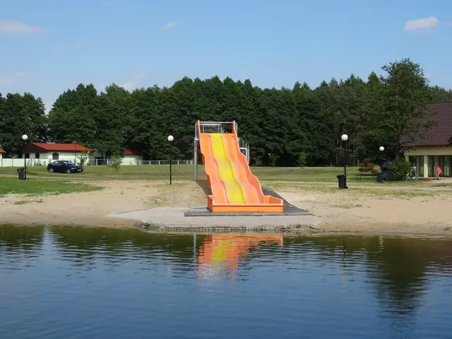 The image shows a playground with a water slide in the middle of a lake surrounded by sand, light...