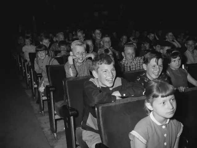The image shows a black and white photo of a group of children sitting in a movie theater, with a...