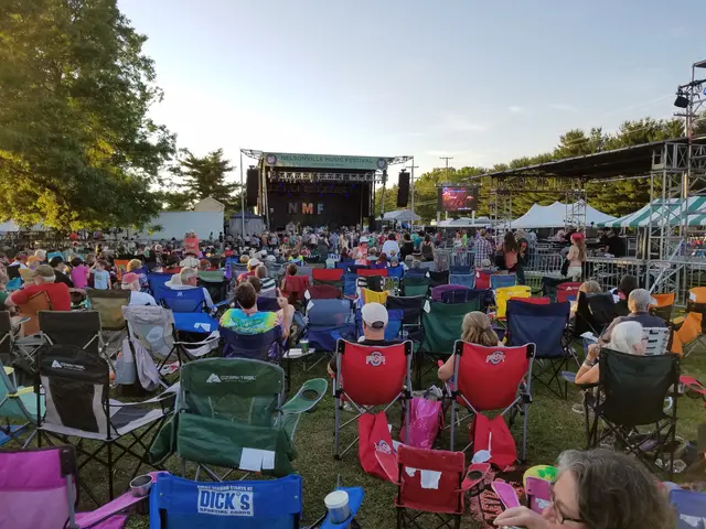 The image shows a large crowd of people sitting in lawn chairs in front of a stage at the...