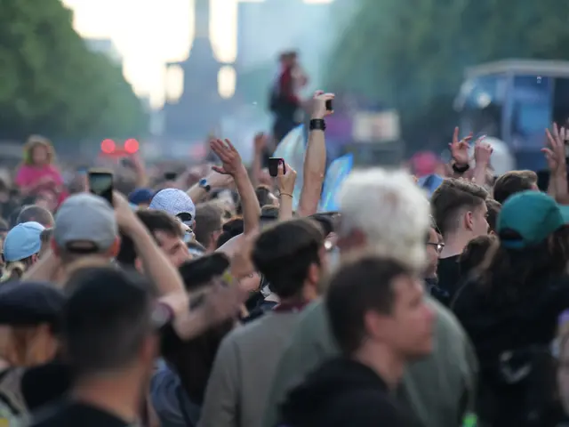 The image shows a crowd of people at a music festival with their hands in the air, some wearing...