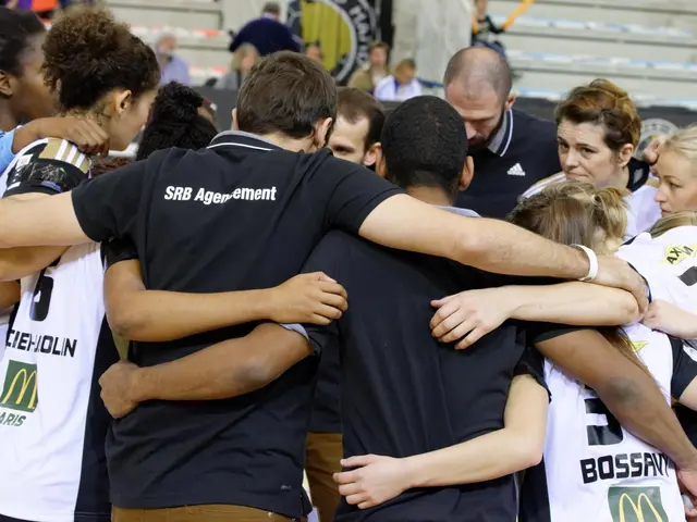 The image shows a group of women's basketball players huddled together in a circle on a court, with...