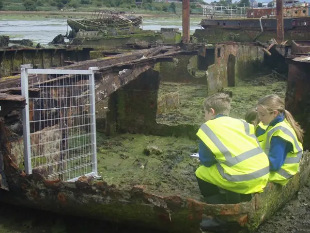 The image shows two children in safety vests sitting on the ground next to a rusty boat, surrounded...
