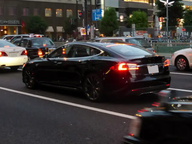 The image shows a black Tesla Model S driving down a city street at night. There are other vehicles...