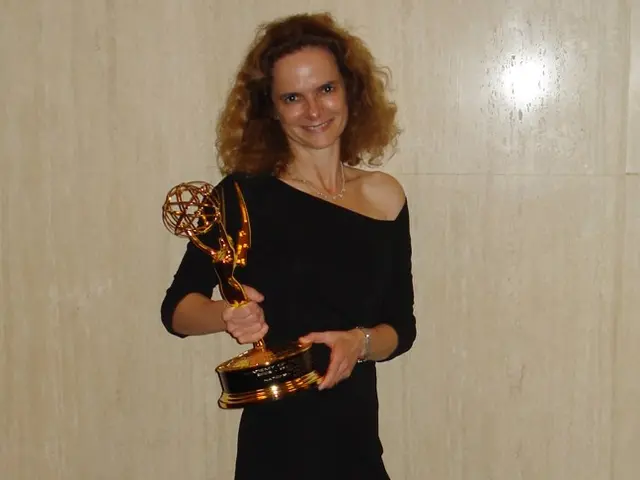 The image shows a woman in a black dress standing on a red carpet, smiling and holding an Emmy...