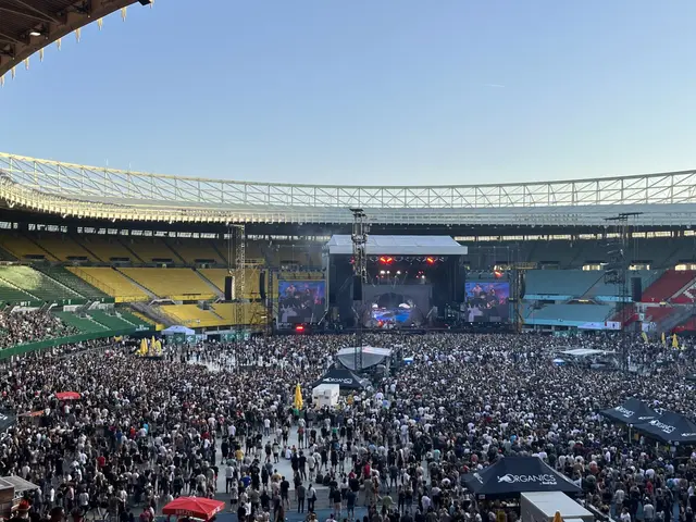 The image shows a large crowd of people standing in front of a stadium, with umbrellas, tents, and...