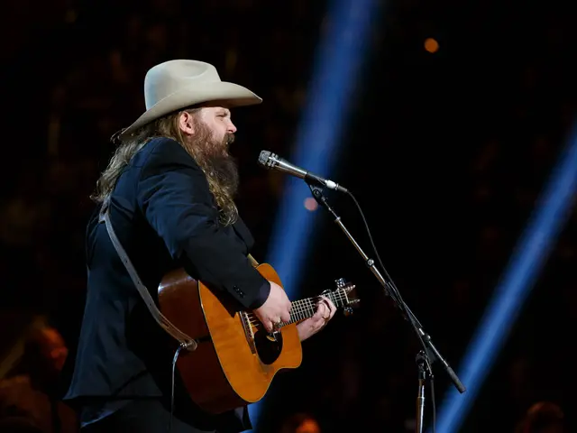 The image shows Chris Stapleton wearing a hat and playing a guitar while singing into a microphone,...