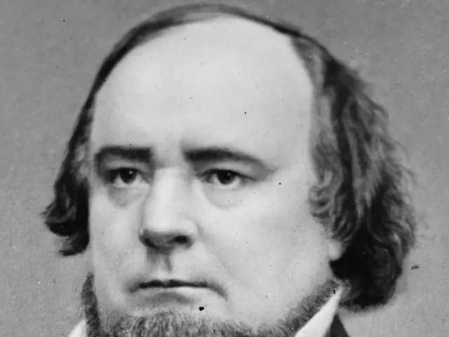 The image shows a black and white photo of a man with a beard wearing a blazer and a bow tie. He is...