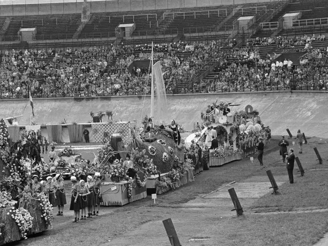 The image shows a black and white photo of a parade in a stadium. There are many people in the...