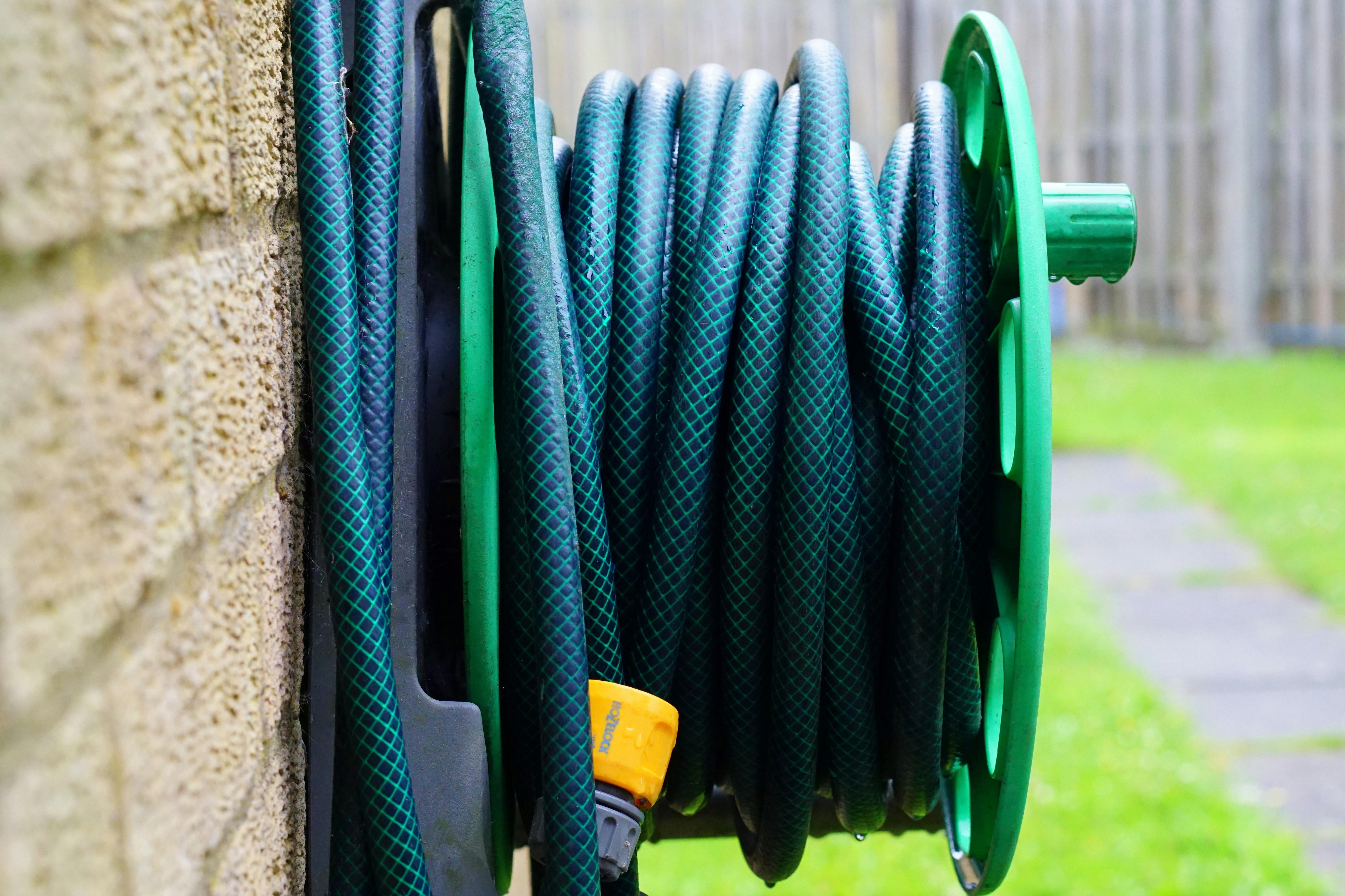 Curled garden hose in a decorative mount, surrounded by fallen autumn leaves on the ground. Decorative garden hose holder holds curled green hose amidst scattered autumn leaves on the floor.