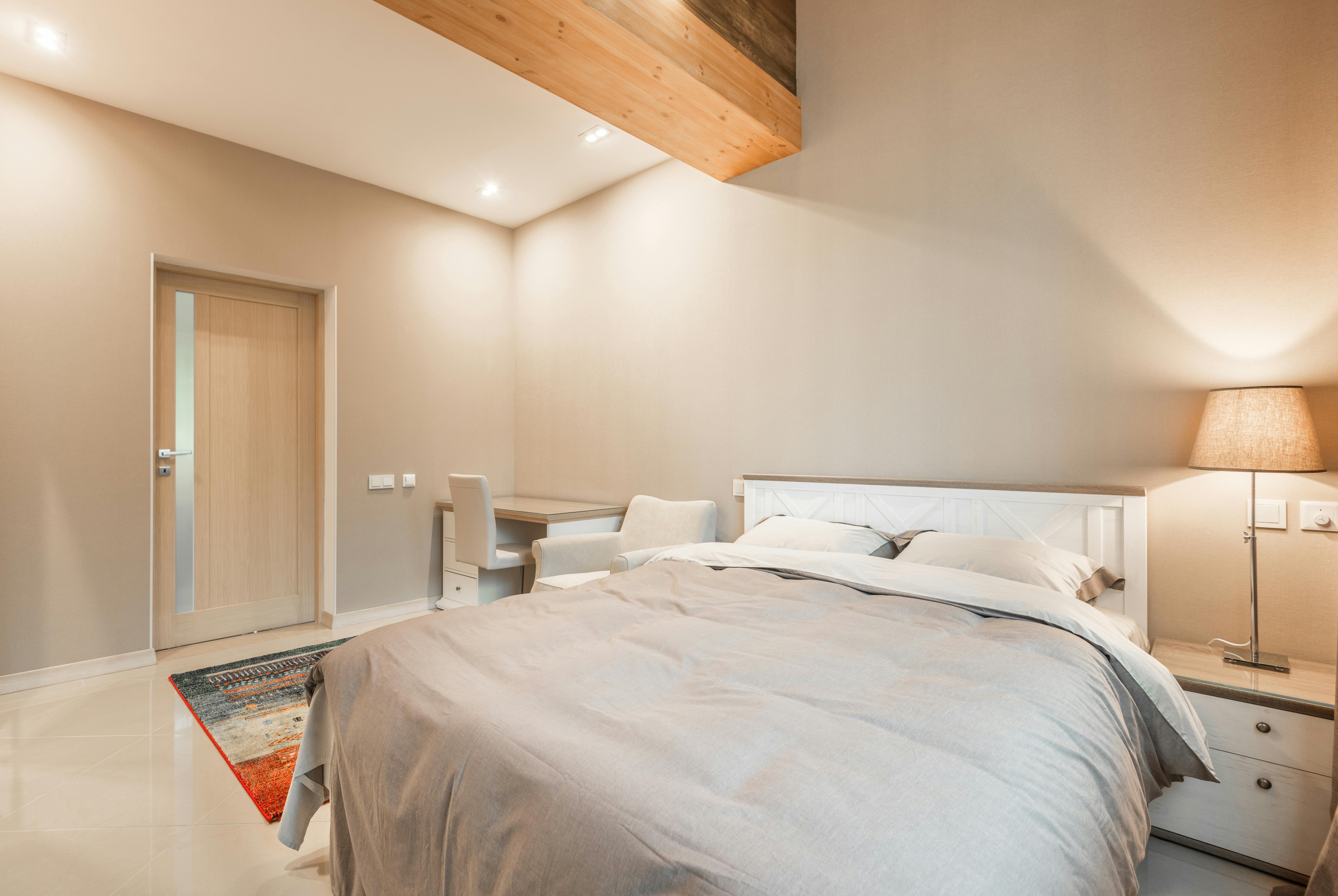 Flower-adorned wall, wooden floor, wicker seating arrangement with cushions, lamp-adorned nightstand, and a white-covered sleeping area. Cozy room decorated with floral wallpaper, a wooden floor, a wicker chair with cushions, a bedside table with a lamp, and a bed draped in white linens.